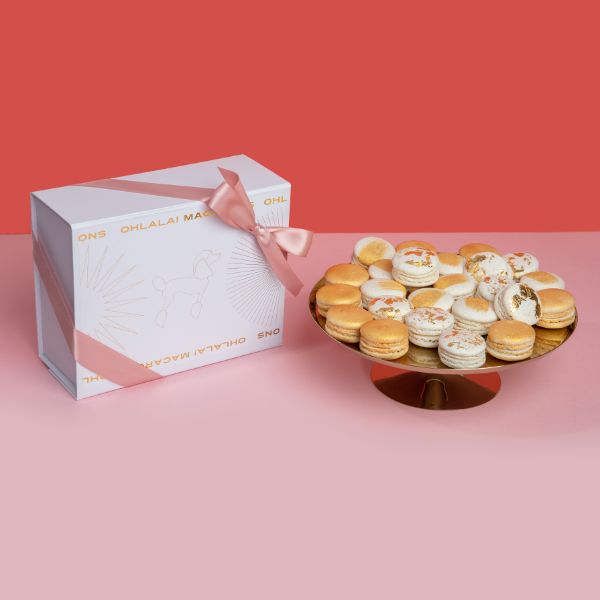 A full plate of macarons sitting next to a white gift box wrapped in a pink bow at Oh La La! Macarons