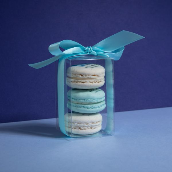 Two white branded macarons and a blue branded macaron in a clear container with a blue bow