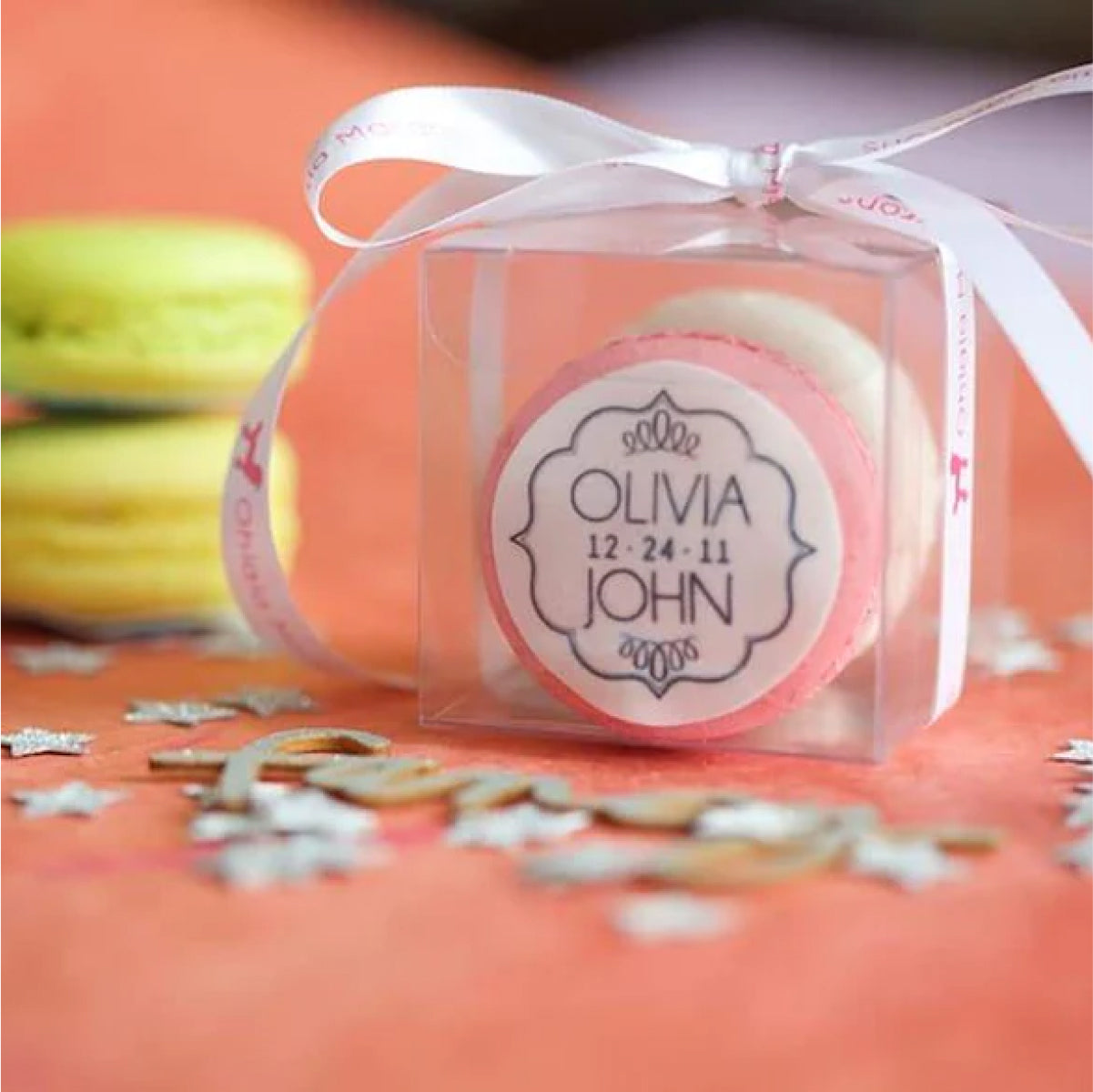 A pink wedding macaron displayed in a clear box that reads "Olivia / 12 - 24 - 11 / John"